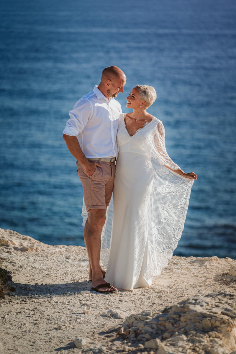 Husband and wife gaze at each other while the bride holds her train to the wind on the Cyprus coast