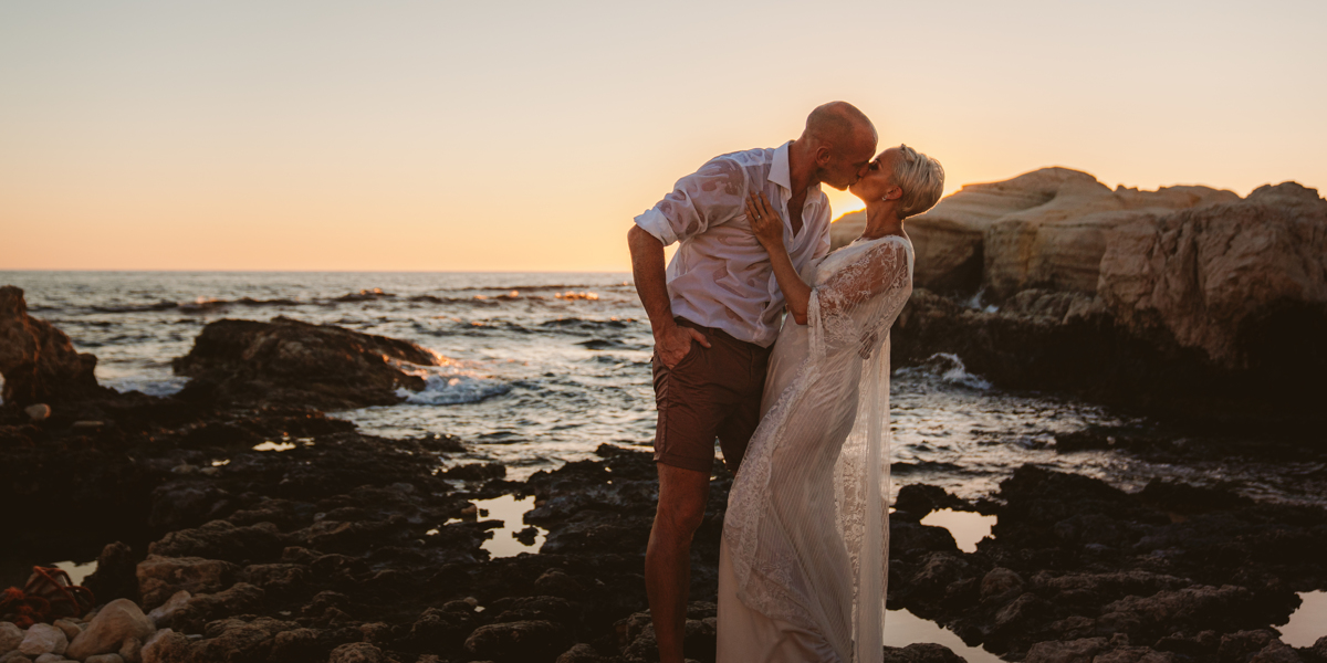 Newlyweds kiss and dip as the sun sets behind them, captured by their Cyprus sea caves wedding photographer